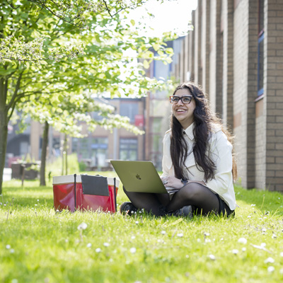 A young female with long dark hair is sitting on the grass, smiling, using a laptop