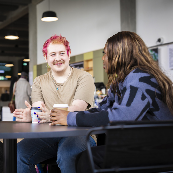 Three casually dressed students are sitting at a table in the student cafeteria, they are chatting and drinking coffee