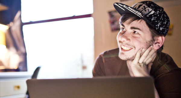A male student wearing a cap is smiling and using his laptop