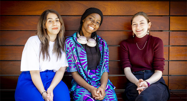 Three female students are smiling and casually sitting outside a university campus building