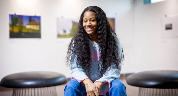 A young female with long dark hair is sitting in an art and design space, smiling for a photo