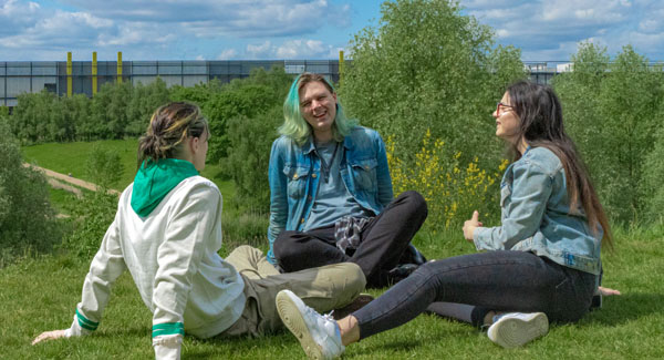 A group of casually dressed students are sitting on grass chatting near the London campus