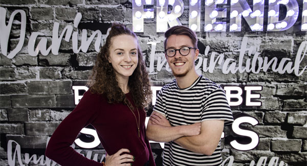 A young female and male are smiling in front of a grey wall with positive wording