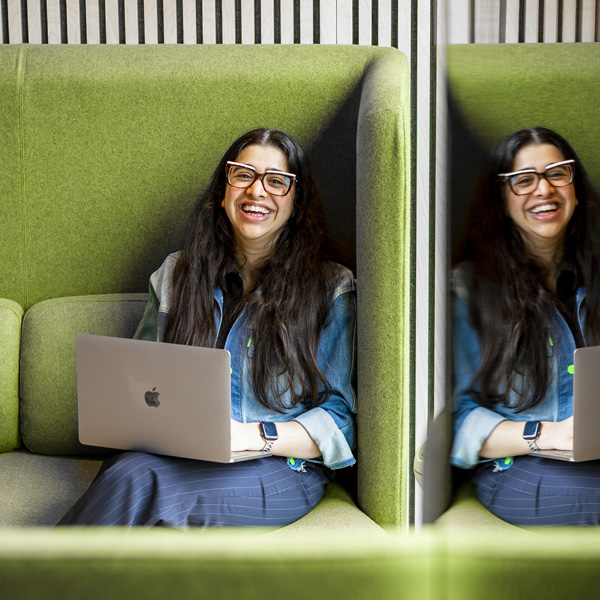 A female with long dark hair is smiling using a macbook whilst sitting on a green office sofa