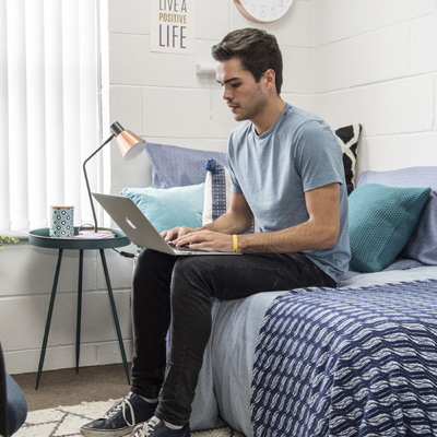A young male wearing a blue tshirt is using his laptop in student accommodation